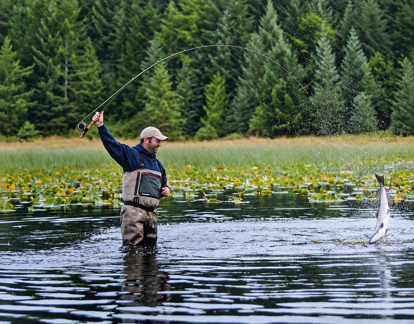 An angler with a bent fly rod as a fish leaps from the water beside a forested shore